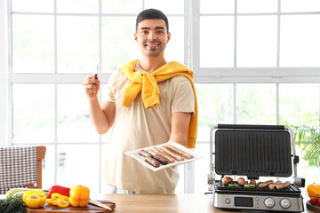 Young man with plate of tasty sausages cooked on modern electric grill at table in kitchen