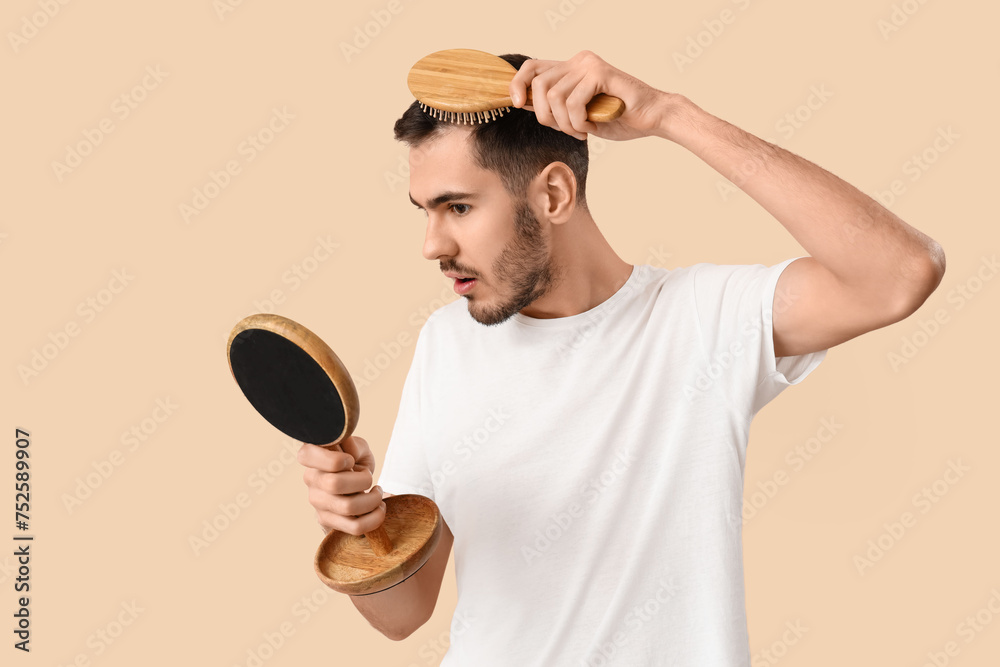 Young man with hair loss problem and brush looking in mirror on beige background