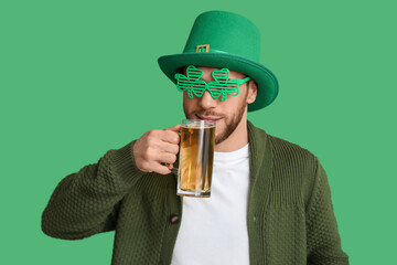 Young man in leprechaun hat and decorative glasses in shape of clover with glass of beer on green background. St. Patrick's Day celebration