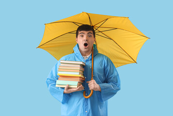 Wall Mural - Shocked young man in raincoat with umbrella and books on blue background