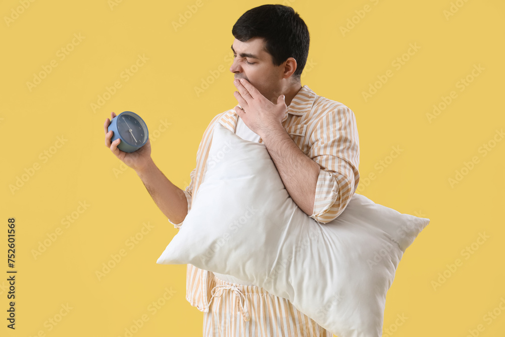 Sleepy young man with alarm clock and pillow on yellow background