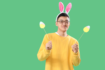 Sticker - Young Asian man in bunny ears with paper eggs on green background. Easter Sale