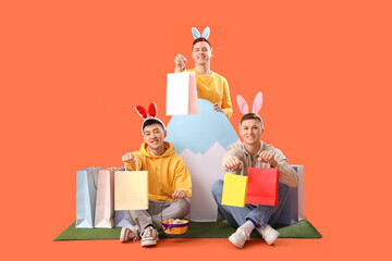 Wall Mural - Young men in bunny ears with shopping bags and paper egg on grass against orange background. Easter Sale