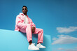 © Brastock Images / AI - Cool young black man in pink attire posing sitting at a blue block under the blue sky. Generative AI