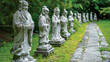 A line of stone statues depicting various traditional Chinese herbalists p along a sacred path in a serene garden.
