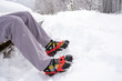 Male legs of a man sitting on a snow covered bench.  The man is wearing gray trousers and mountain boots with Grödel on them.  Grödel, a type of spikes, are used for winter hikes on icy surfaces.