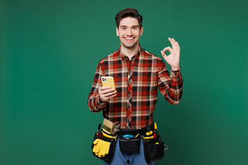 Wall Mural - Young employee laborer handyman man wear red shirt hold use mobile cell phone show ok okay isolated on plain green background Instruments accessories for renovation apartment room Repair home concept