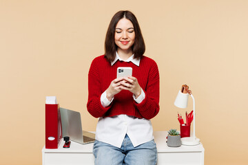 Wall Mural - Young employee business woman wear red sweater shirt work at office sit on desk with pc laptop hold in hand use mobile cell phone isolated on plain pastel beige background. Achievement career concept