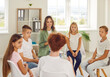 Friendly counselor is talking to group of junior high school students at psychological session. Smiling woman sits in classroom in circle with group of children and listens to schoolboy's story.