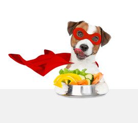 Poster - Licking lips Jack russell terrier puppy wearing superhero costume holds bowl of vegetables above empty white banner. Isolated on white background