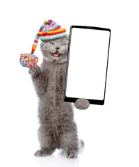 Poster - Happy cat wearing warm knitted hat with pompon and woolen scarf holding smartphone with white blank screen in it paw. Isolated on white background
