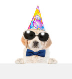 Fototapeta Psy - Golden retriever puppy wearing tie bow, sunglasses and party cap looks above empty white banner. isolated on white background