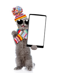 Poster - Happy cat wearing warm knitted hat with pompon and woolen scarf holding smartphone with white blank screen in it paw. Isolated on white background