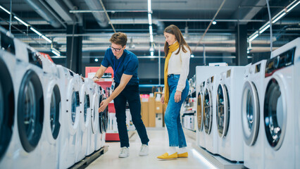 Wall Mural - Customer Engages in Dialogue with Retail Electronics Shop Consultant while Choosing a Washing Machine. Female Searching for a Laundry Appliance. Shopper Exploring Products in a Modern Retail Store