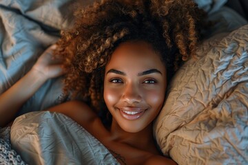 Wall Mural - Cheerful young woman with curly hair covered with a bedsheet in bed, smiling