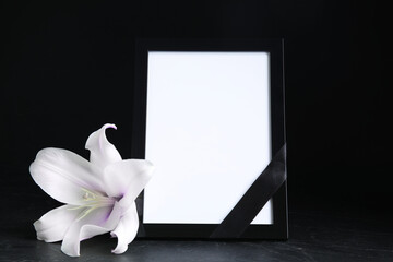 Wall Mural - Funeral photo frame with ribbon, white and violet lily on dark table against black background