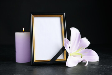 Wall Mural - Funeral photo frame with ribbon, violet lily and candle on dark table against black background