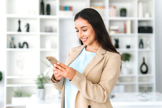 Using gadgets. Happy successful arabian or indian woman in a suit, stands in the office, holding mobile phone, texting in social media with friends or clients, making online order, smiles