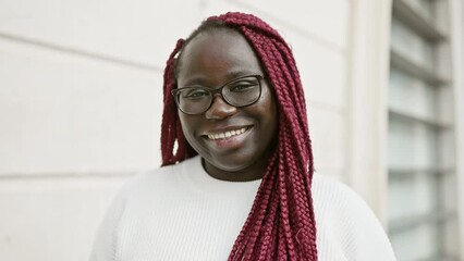 Sticker - A cheerful african woman with burgundy braids and glasses standing on an urban street.