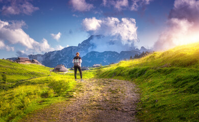 Wall Mural - Girl on the rural road with yellow flowers and green grass in beautiful alpine mountain valley at sunset in summer. Young woman in old alpine village, blue sky with clouds. Travel and Hiking. Slovenia