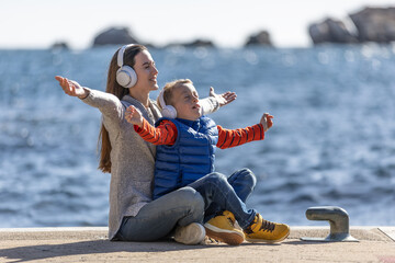 Wall Mural - Mother and son with arms oustretched, sitting on a pier listening to music with headphones, and breating fresh sea air