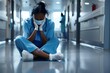 © pham - Tired depressed female African scrub nurse wears face mask blue uniform gloves sits on hospital floor