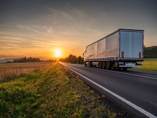 Wall Mural - White truck driving on the asphalt road in rural landscape at sunset