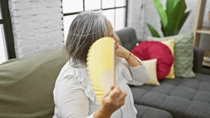Wall Mural - Mature woman cools off with a yellow fan in a cozy, well-decorated living room, exuding elegance and comfort.