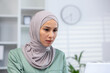 © Liubomir - Focused Muslim woman wearing a hijab experiencing concern or stress while working in a modern office setting.