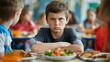 Unhappy young boy with a frown sitting in a cafeteria, his elbows on the table, looking dissatisfied with a healthy lunch plate in front of him