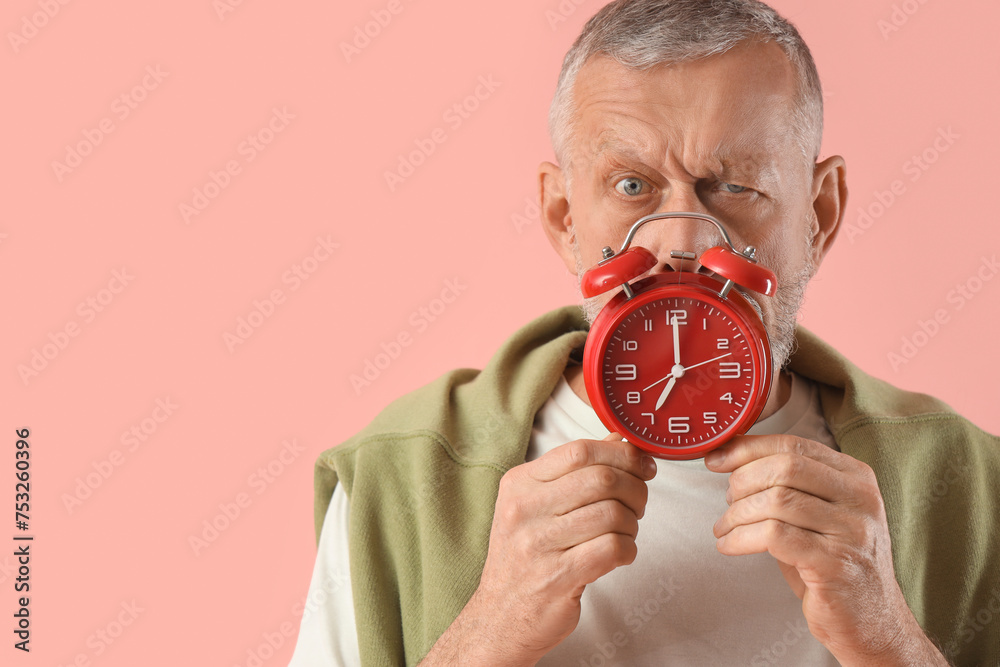 Thoughtful mature man with alarm clock on pink background, closeup