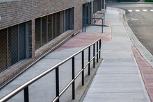 The Exterior Wall Of A Large Brick Building With A New Concrete Sidewalk. There's A Wheelchair Access Ramp. There's A Black Metal Handrail With A Barrier Free Surface. The Path Is Grey And Pink Color.
