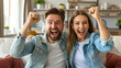 © MVProductions - Excited couple celebrating success while sitting on sofa at home. Overjoyed happy young man and woman in casual wear raising clenched fists in excitement doing winner gesture