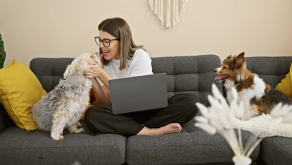 Canvas Print - A hispanic woman interacts with her dogs on a couch in a cozy living room while using a laptop.