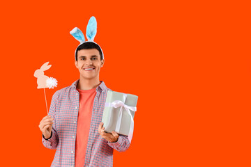 Poster - Happy young man in bunny ears with gift box and paper rabbit on orange background. Easter celebration