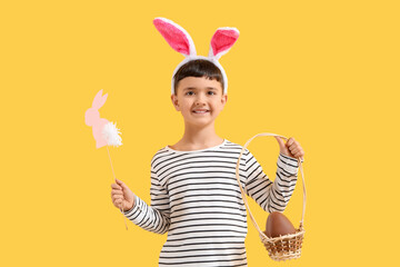 Sticker - Cute little boy in bunny ears headband holding basket with chocolate egg and decor for Easter on yellow background