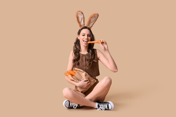 Wall Mural - Happy young woman in Easter bunny ears headband with paper shopping bag eating carrot on beige background