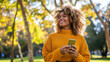 A young woman smiles as she holds a smartphone in her hands, glancing away while enjoying the outdoors in the park.