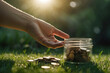 Cropped hands and coins in a glass jar on the green grass with the morning sun in the background creating a flare