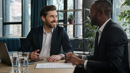 Two diverse businessmen Caucasian man and African American businessman with laptop computer in office corporation company talking discuss online result business strategy marketing planning laughing