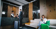 © ihorvsn - Gorgeous businessman checking in hotel room having conversation with professional and kind receptionist helping guests. Beautiful young lady relaxing on sofa in contemporary hotel.
