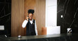 © ihorvsn - Portrait of lovely elegant professional female receptionist using phone to speak with clients and looking at camera smiling. Attractive African-American administrator of modern hotel.