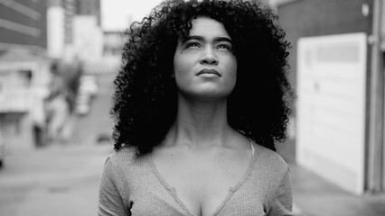 One hopeful young black woman with curly hair standing in street looking up at sky with HOPE and FAITH in black and white. African American person feeling the presence of GOD