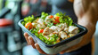 Man holding a plastic black container box with healthy fitness meal including white meat, fresh green salad and vegetables. Tasty lunch with high protein foods, for muscle building, gym interior