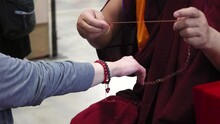 The monk dressed in traditional attire is tying a red string around another person's wrist. It is a religious ceremony that holds special significance in the culture or tradition by the monk.