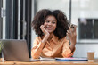 © Songsak C - Portrait of a beautiful confident businesswoman using a laptop computer holding a mobile phone sitting in a modern office. Smiling African American freelancer working online from home.