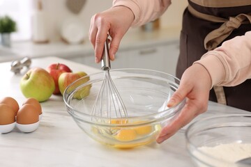 Wall Mural - Woman whisking eggs in bowl at light marble table indoors, closeup