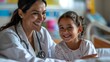 A smiling female doctor with a stethoscope around her neck sitting beside a young girl patient on a hospital bed.