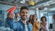 The happy male leader of the company threw a red paper airplane into the office. There was a group of employees smiling and talking in the background.