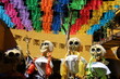 4 catrinas, esqueletos amarillos vestidos de mujeres, con trenzas, en un patio en Oaxaca, México, el día de muertos, con adornos de banderas de colores de papel picado y una pared amarilla.